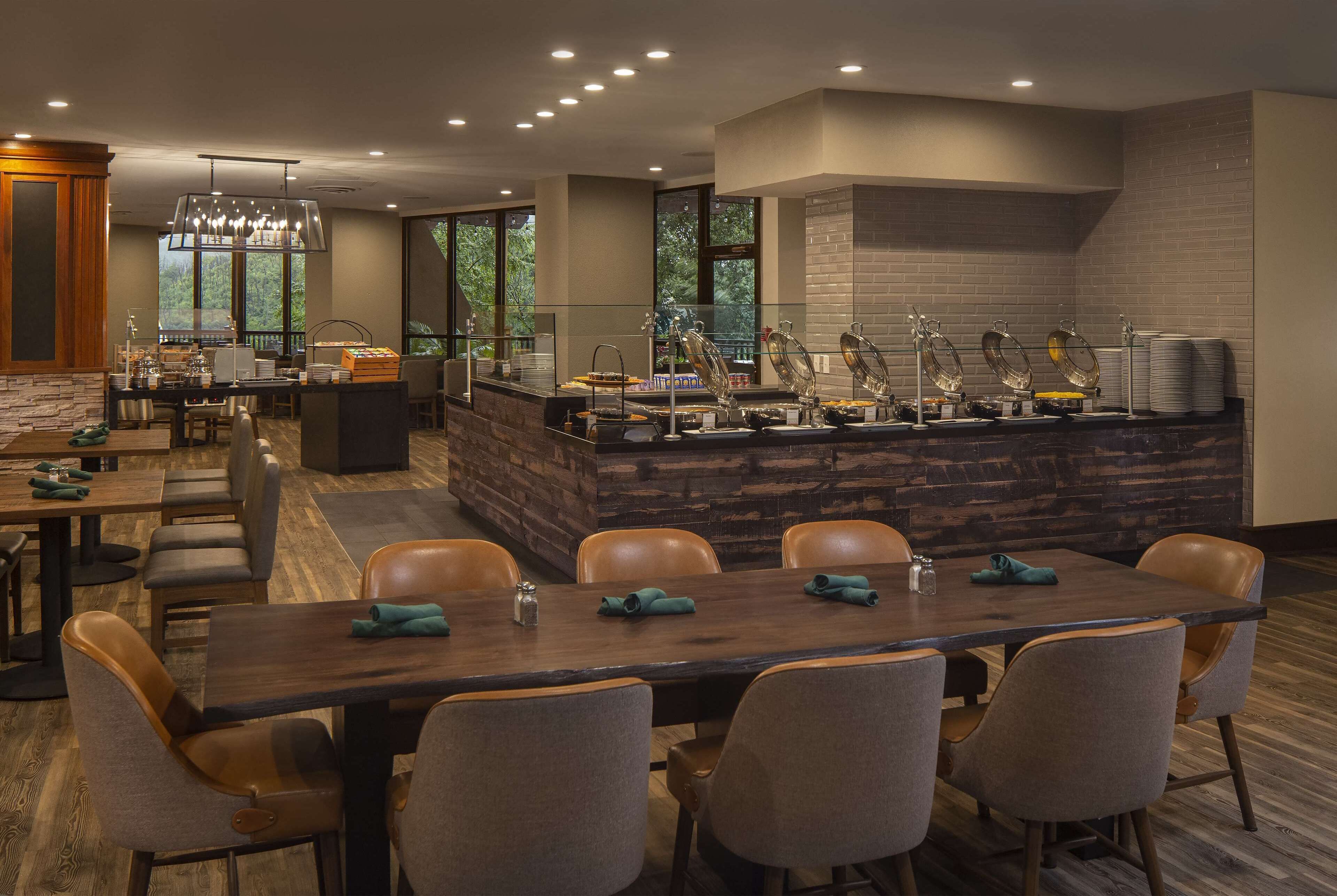 A modern dining area with a long wooden table, cushioned chairs, and a buffet counter with various food items and utensils.