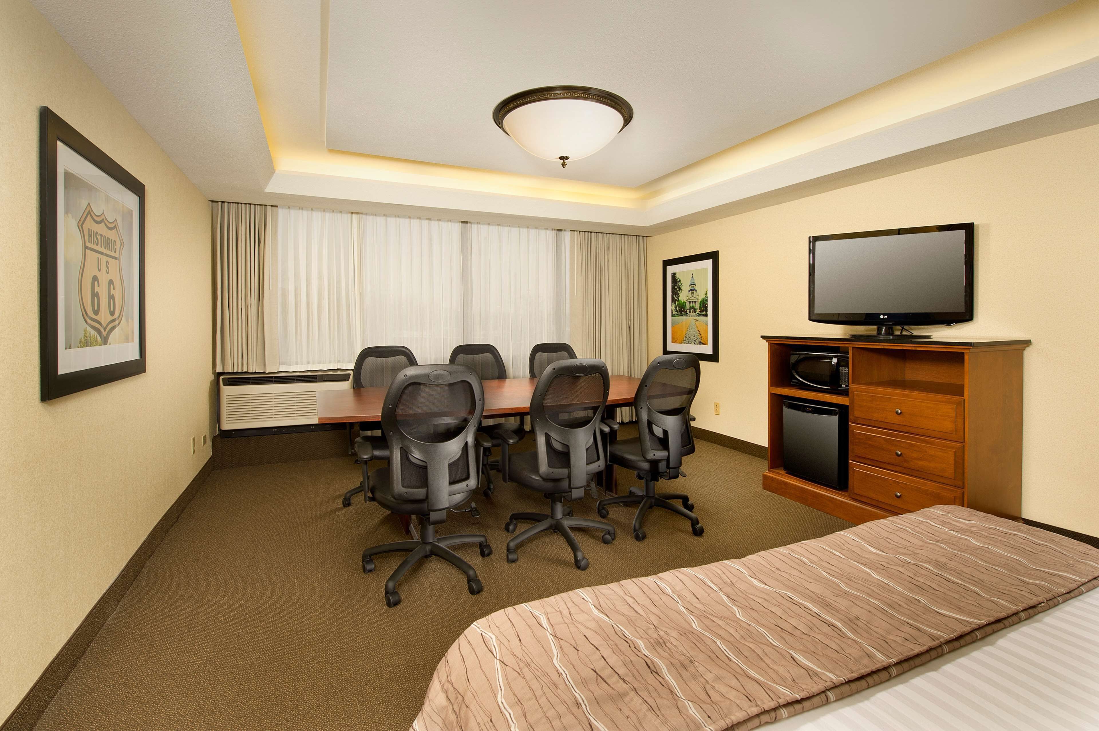 A hotel room with a conference table, four office chairs, a television, a bed, and a small cabinet.