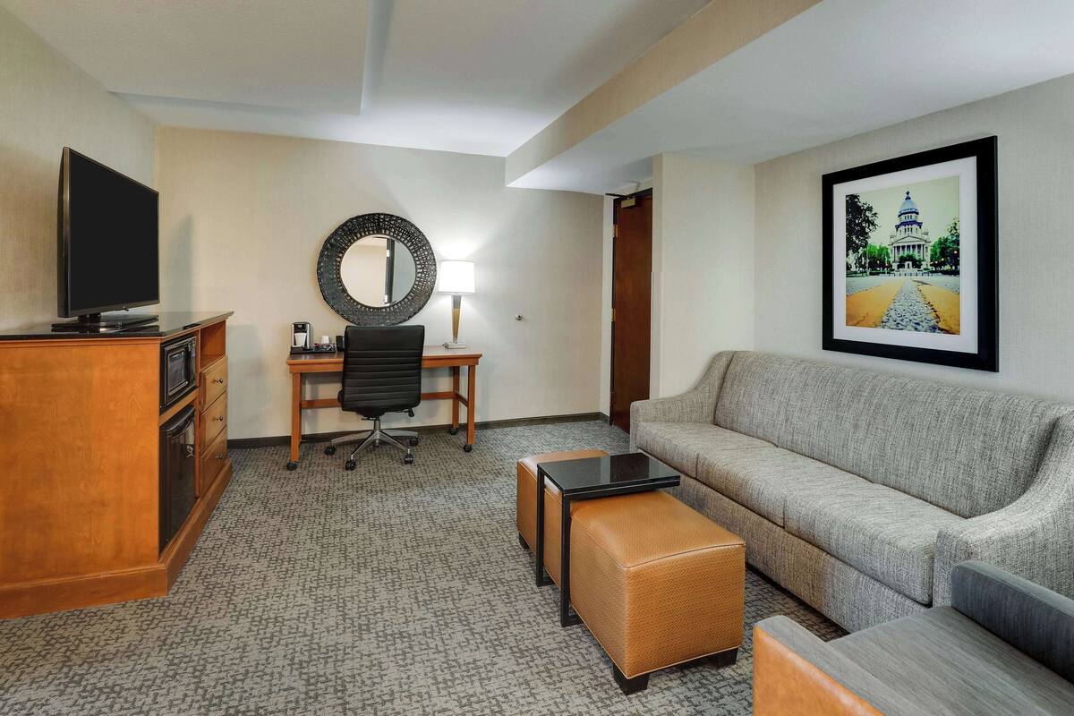 A hotel room with a flat-screen TV, a desk with a chair, a sofa, a coffee table, and a framed picture on the wall.