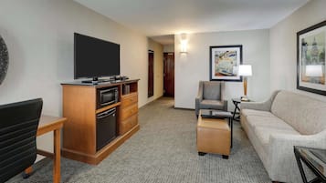 A hotel room with a flat-screen TV, a wooden entertainment unit, a sofa, a chair, and a small table.