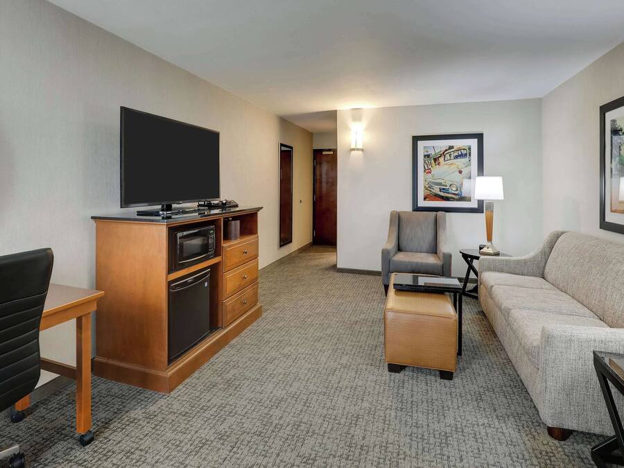 A hotel room with a flat-screen TV, a wooden entertainment unit, a sofa, a chair, and a small table.