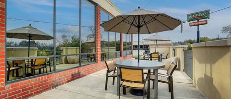 An outdoor patio with a brick wall, large windows, and a table with chairs and umbrellas.