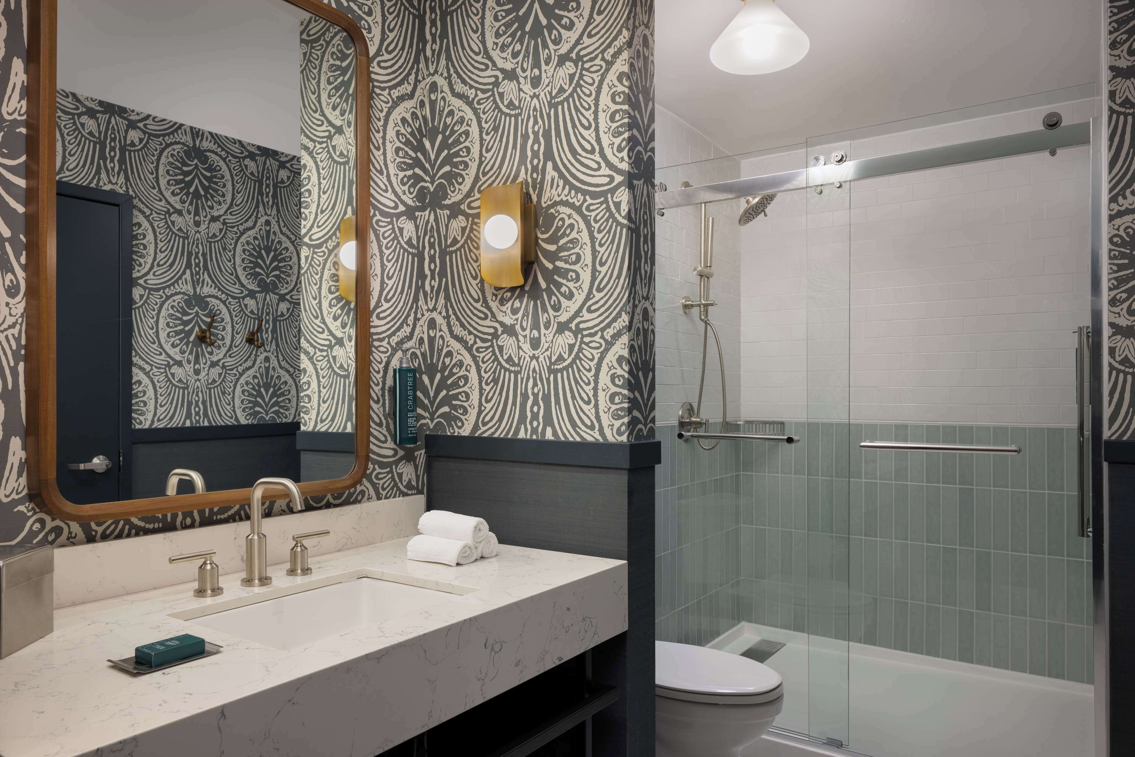 Un baño moderno con pared estampada, un espejo grande, un lavamanos con dos grifos, una toalla y un área de ducha con mampara de vidrio.