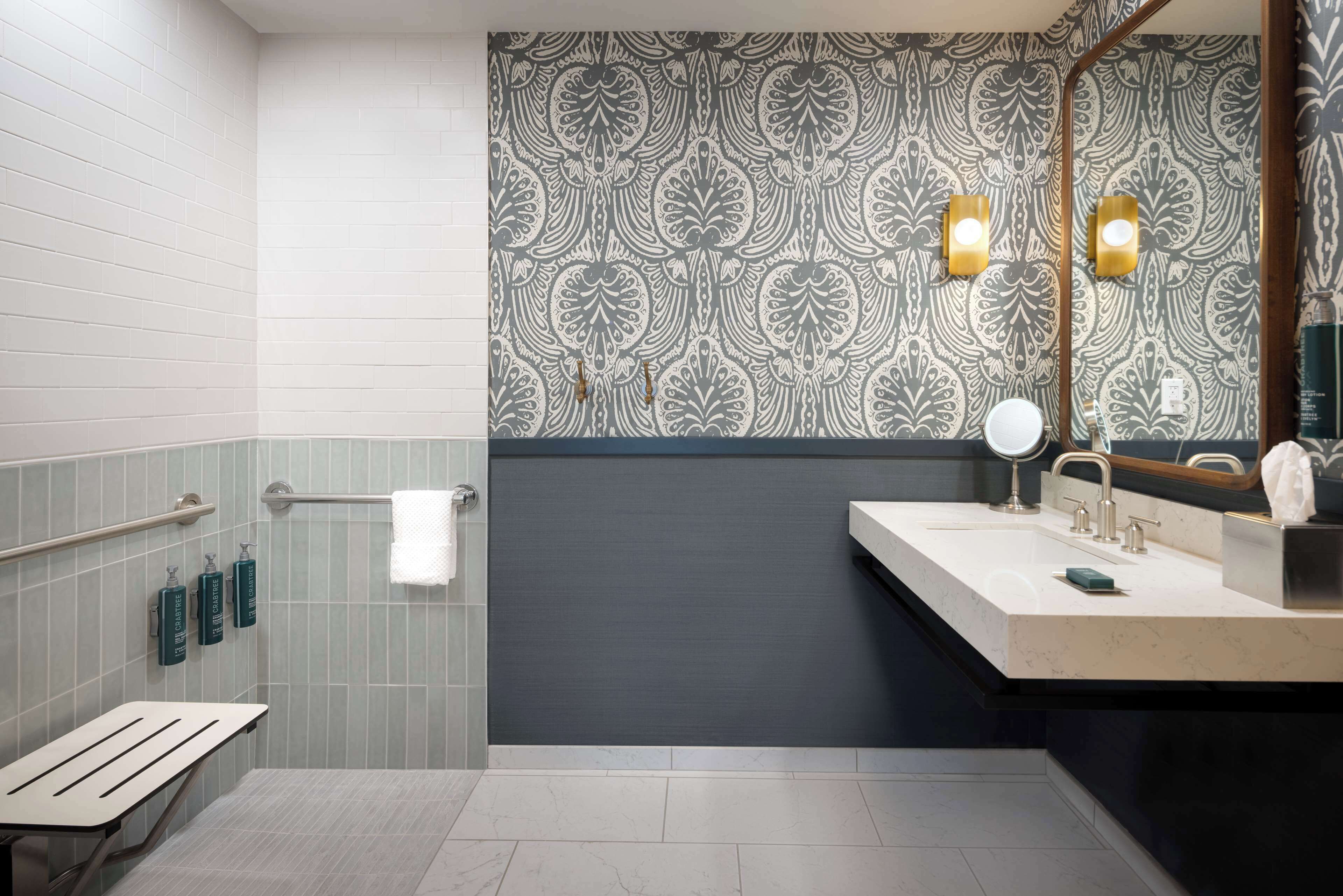 A modern bathroom with a grey and white patterned wall, a dark accent wall, a vanity with a mirror, and a bench.