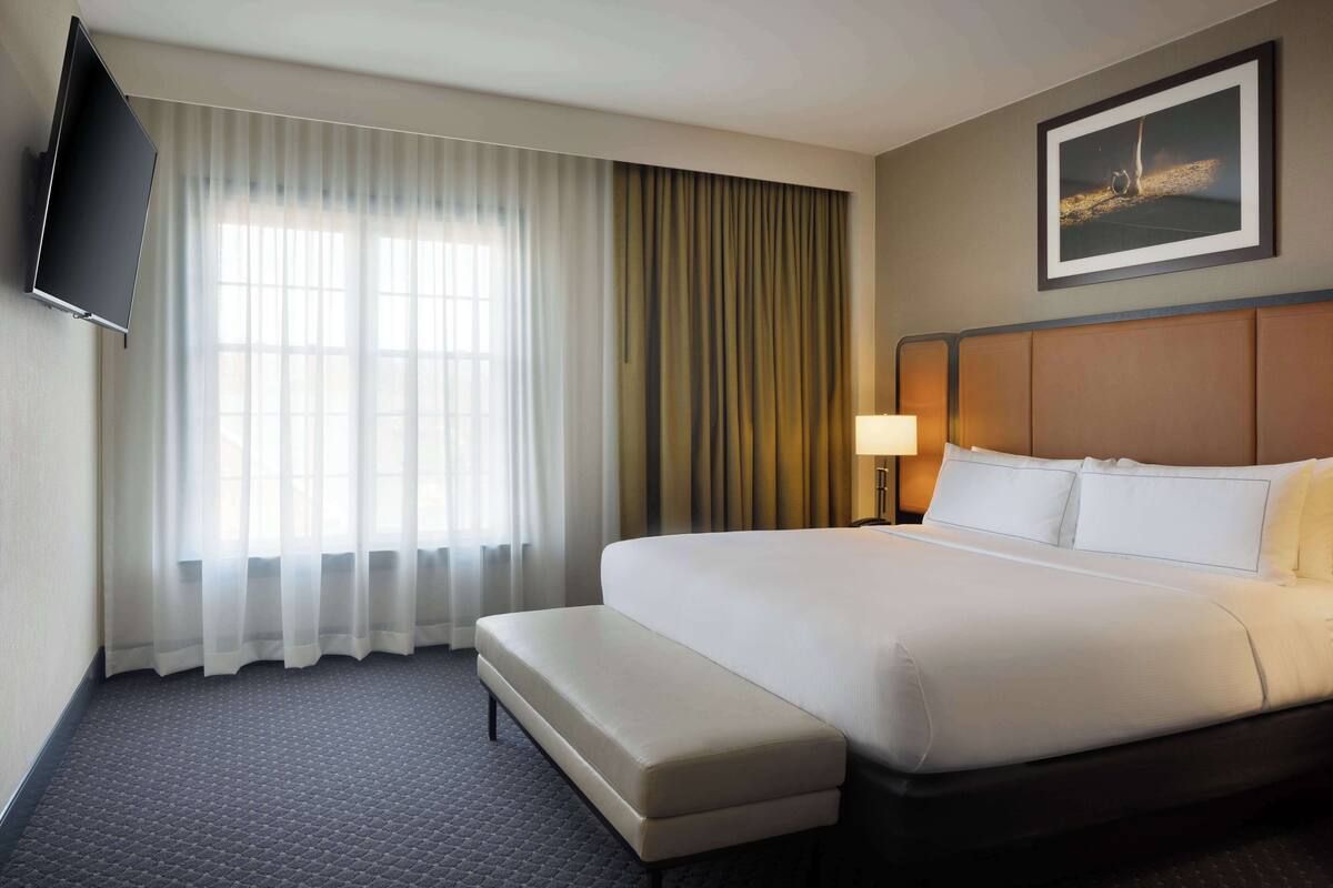 A hotel room with a large bed, a bench, a flat-screen TV mounted on the wall, a window with sheer curtains, and a framed picture on the wall.