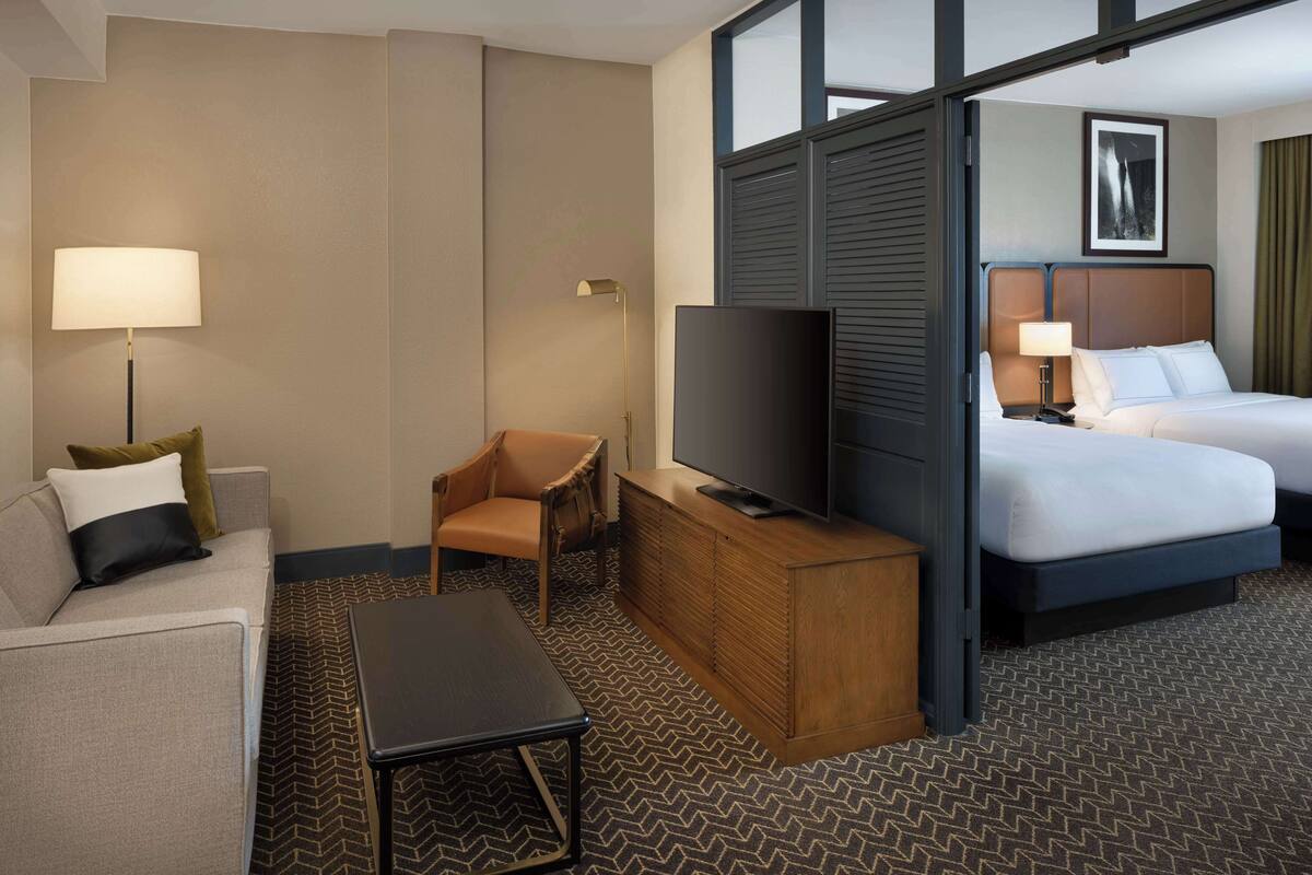 A hotel room with a sofa, a chair, a TV on a wooden cabinet, a bed, and a large window.