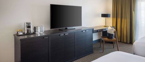 Habitación de hotel con una cama, un televisor de pantalla plana sobre un mueble oscuro, una cafetera y una silla con un cojín.