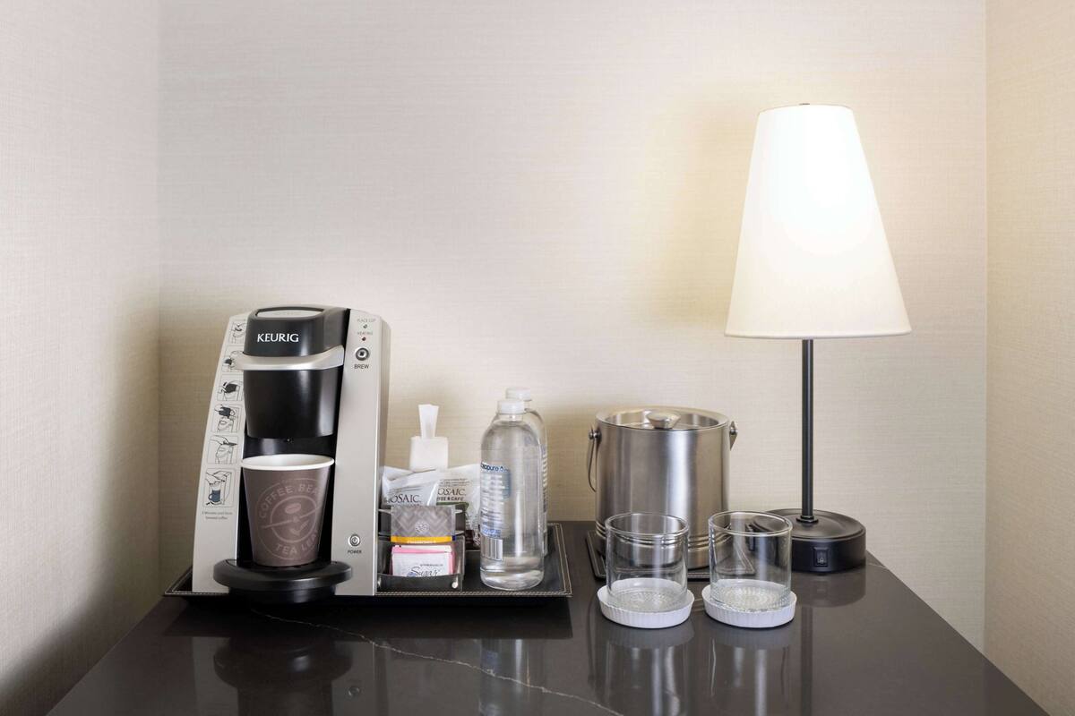 A hotel room with a coffee maker, a water bottle, a glass, a lamp, and a cup on a table.
