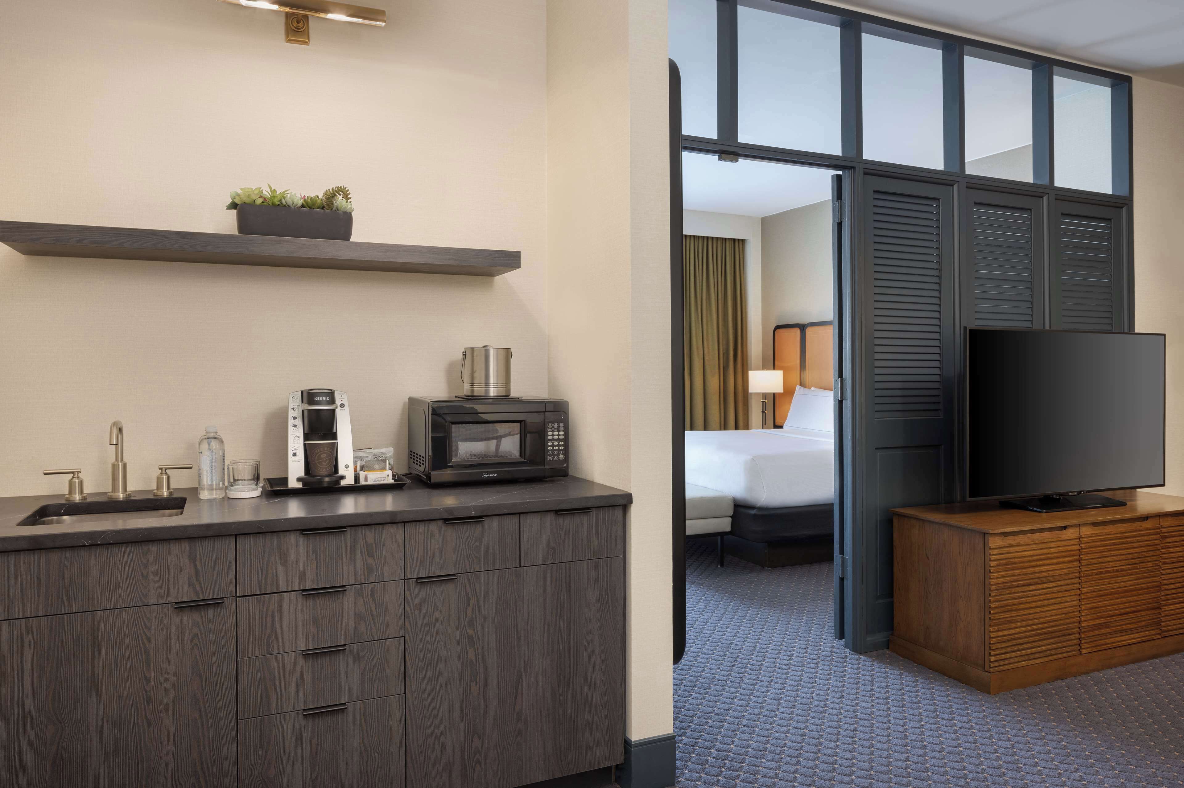 A hotel room with a kitchenette, a flat-screen TV, a bed, and a view through a sliding door.