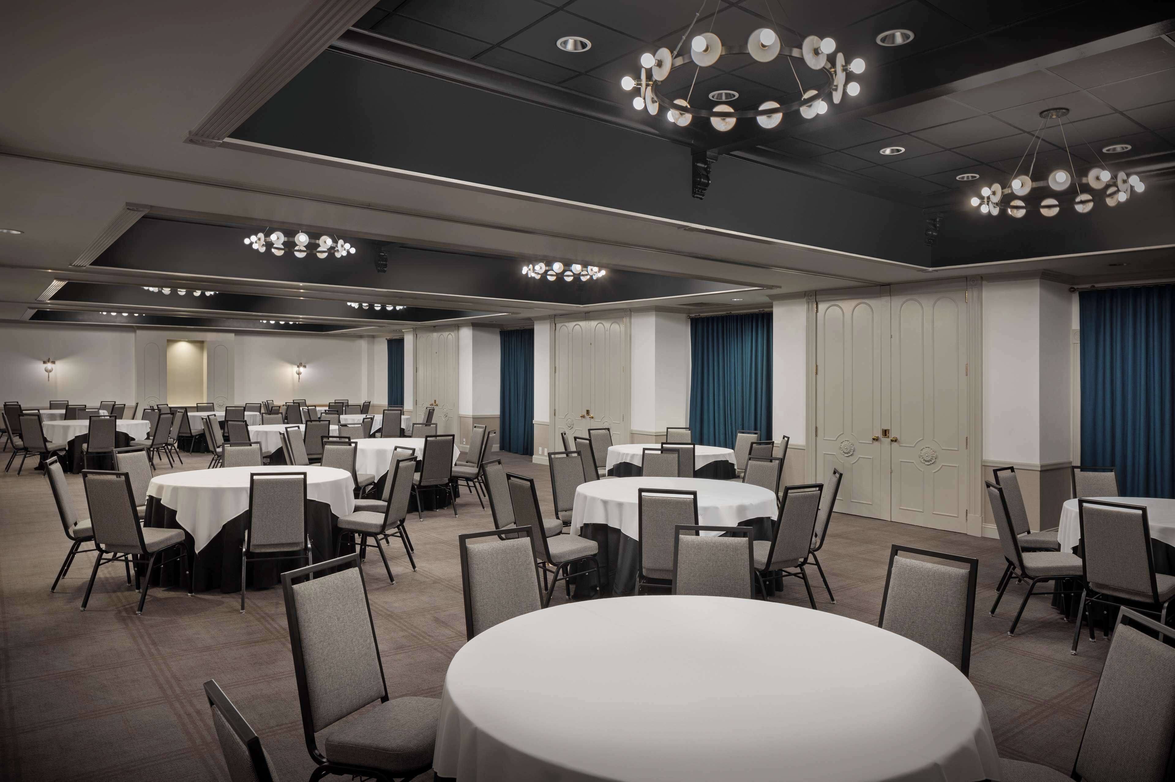 Une salle de conférence dotée de plusieurs tables rondes et de chaises, d’un éclairage au plafond et d’une porte ornée d’un rideau bleu.