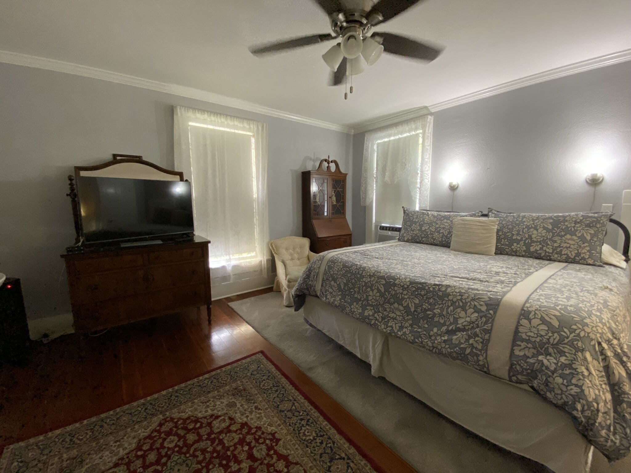 Une chambre à coucher comprenant un lit, une commode, un téléviseur, un ventilateur de plafond et une fenêtre avec des rideaux.