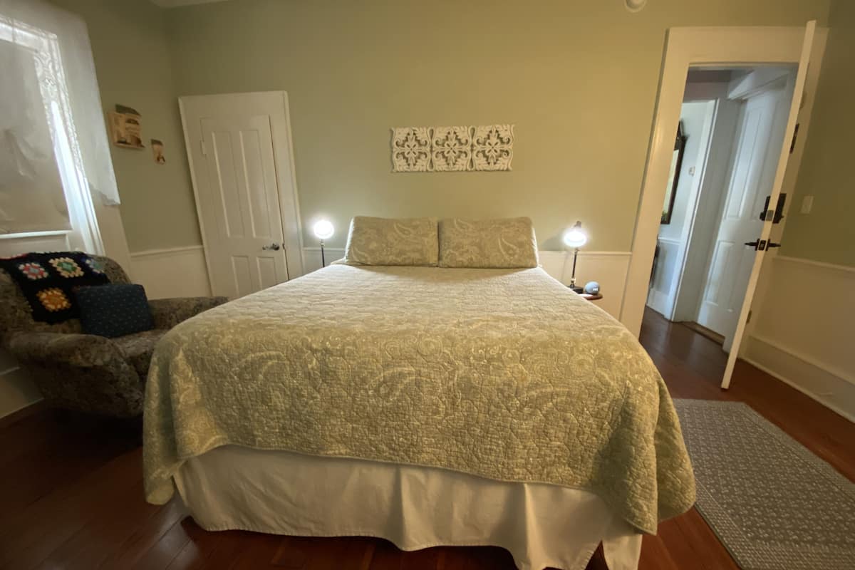 Une chambre avec un grand lit, une table de chevet avec une lampe, une chaise avec une couverture, et une porte donnant sur une autre pièce.