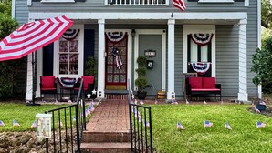 Une maison à deux étages avec un porche à l’avant, décoré de drapeaux américains et de guirlandes aux couleurs patriotiques. On y trouve deux chaises rouges, un petit marchepied menant à l’entrée et une boîte aux lettres portant le numéro 221.