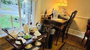 Un coin repas avec une table en bois, dressée avec des tasses à thé, une théière et un bol de collations. Une fenêtre offre une vue sur la rue et la verdure environnante.