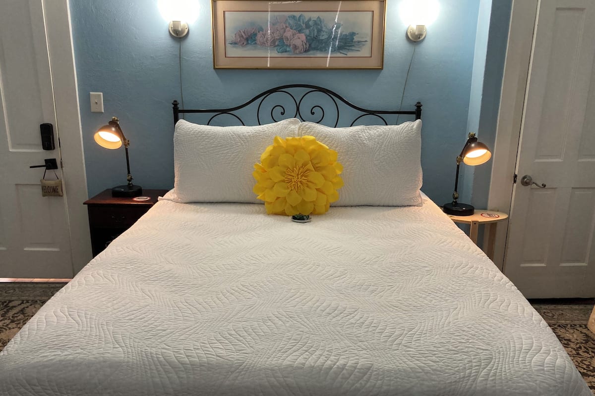 Un lit bien fait, agrémenté d’un grand bouquet de fleurs jaunes, deux lampes de chevet, une œuvre d’art encadrée au mur et une porte sur laquelle est apposée une pancarte « Bienvenue ».