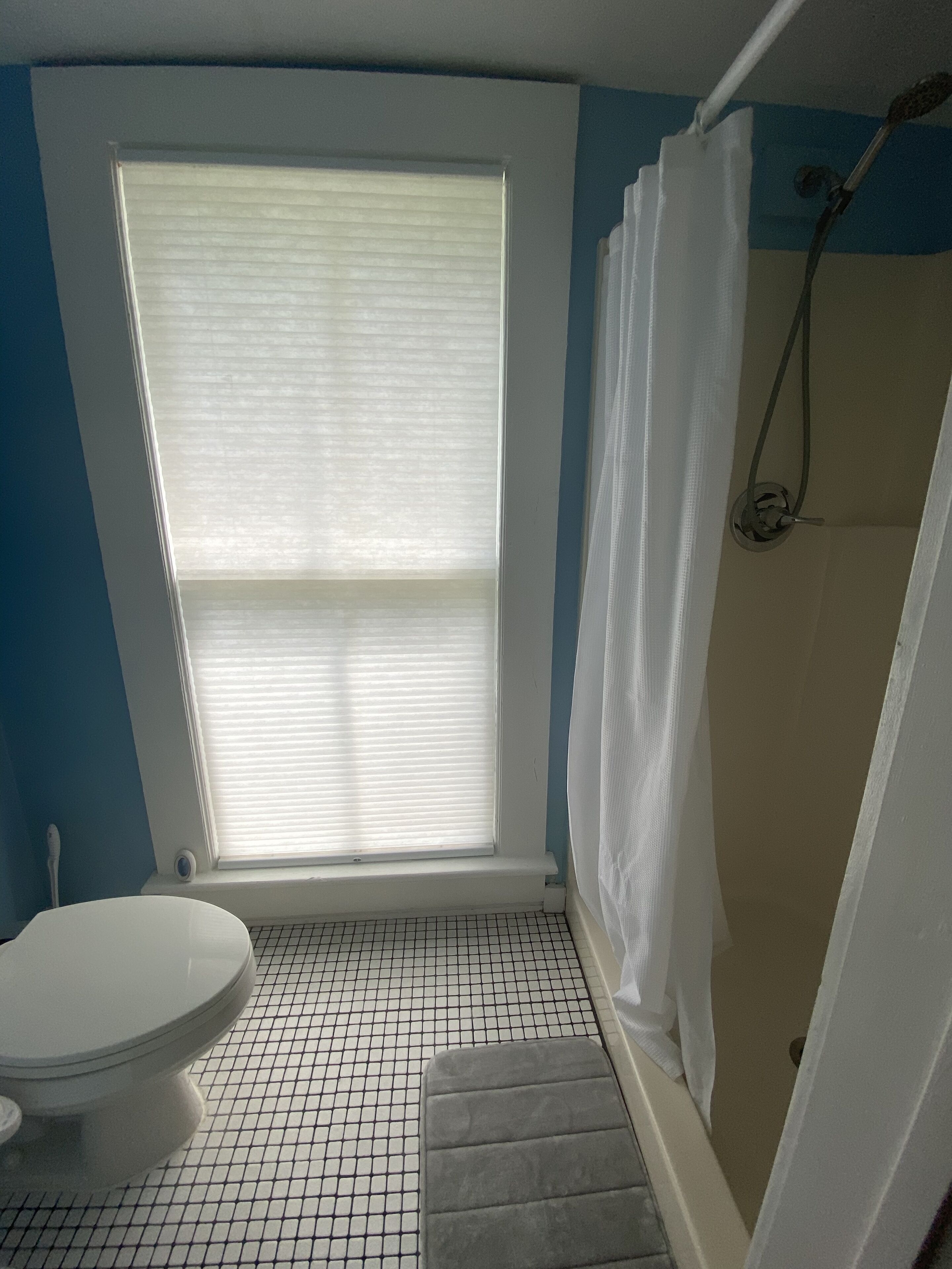 A bathroom with a toilet, a window with blinds, a shower area, and a tiled floor.