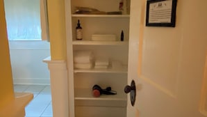 Un couloir mène à une pièce dont la porte est blanche, où l’on peut voir un certificat encadré accroché au mur et une étagère avec des serviettes pliées.