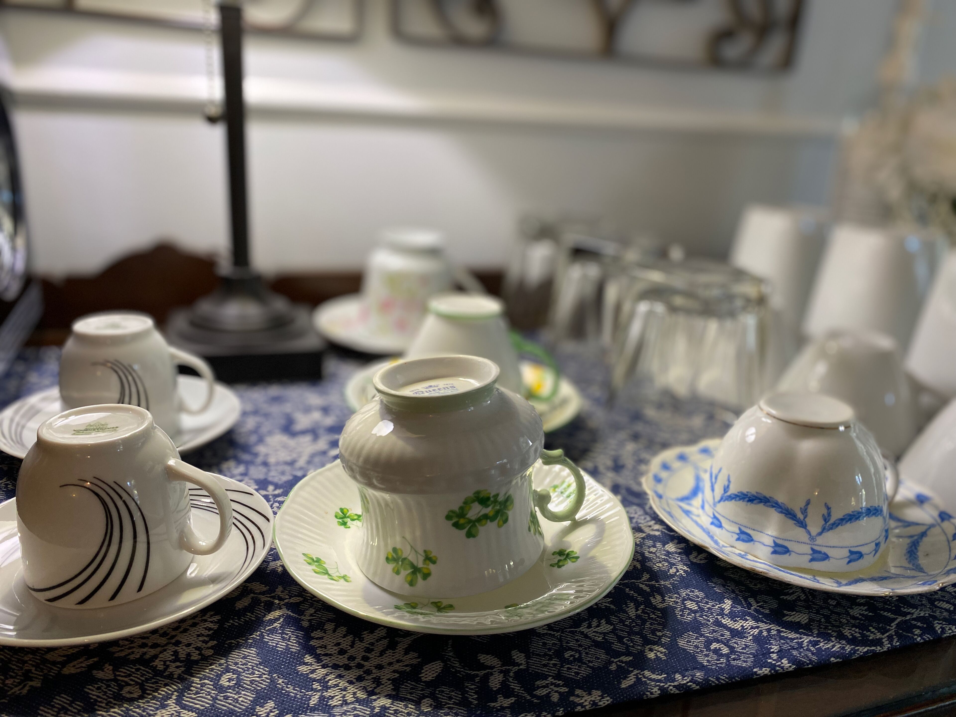 Una mesa con varias tazas y platillos de cerámica blanca, algunos con motivos florales, sobre un mantel azul estampado.