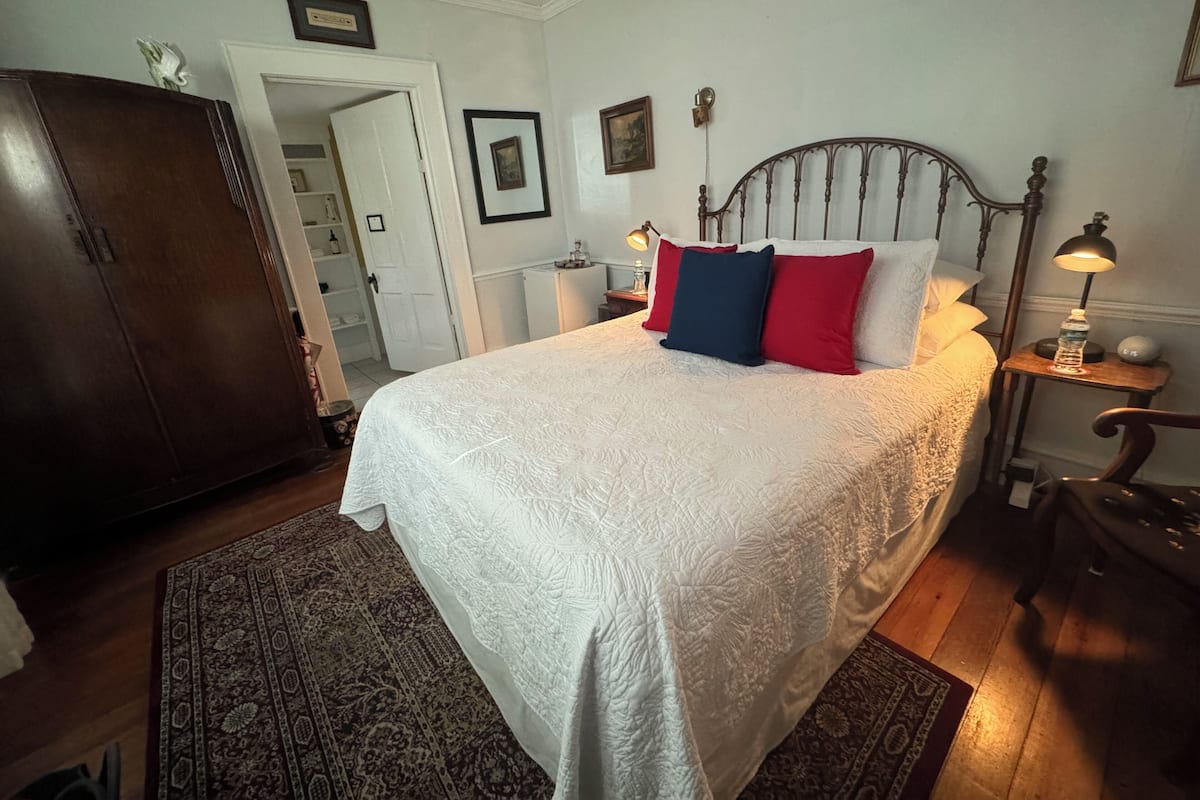 Un lit bien fait, recouvert d’une couette blanche, avec une tête de lit en bois et trois oreillers aux couleurs rouge, bleu et blanc. Une table de chevet avec une lampe et une bouteille d’eau se trouve à côté du lit. On aperçoit une armoire en bois sur le côté gauche, et un tapis à motifs recouvre le sol.