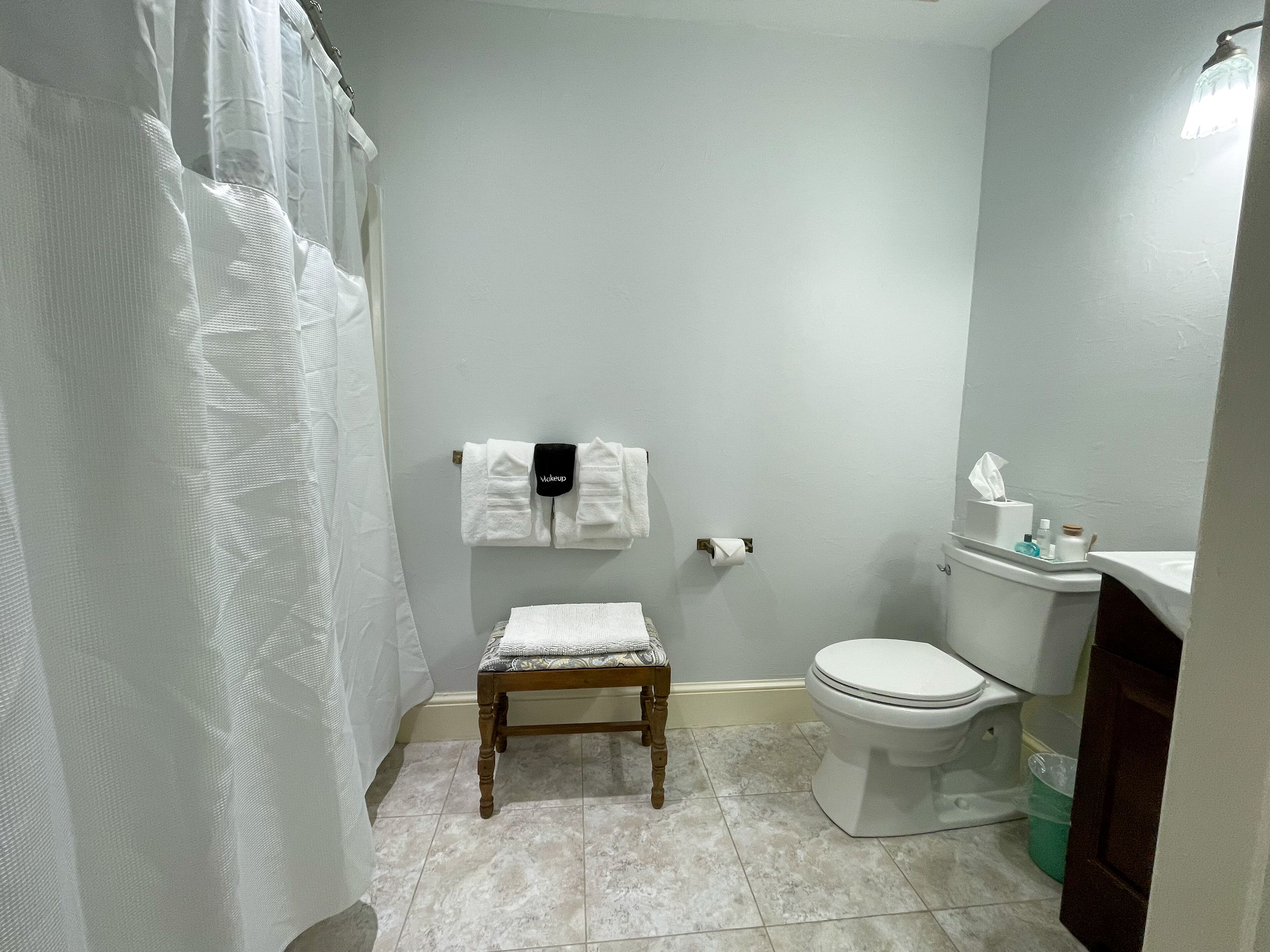 Un baño con cortina de ducha, un taburete de madera, un inodoro y un perchero con una toalla blanca.