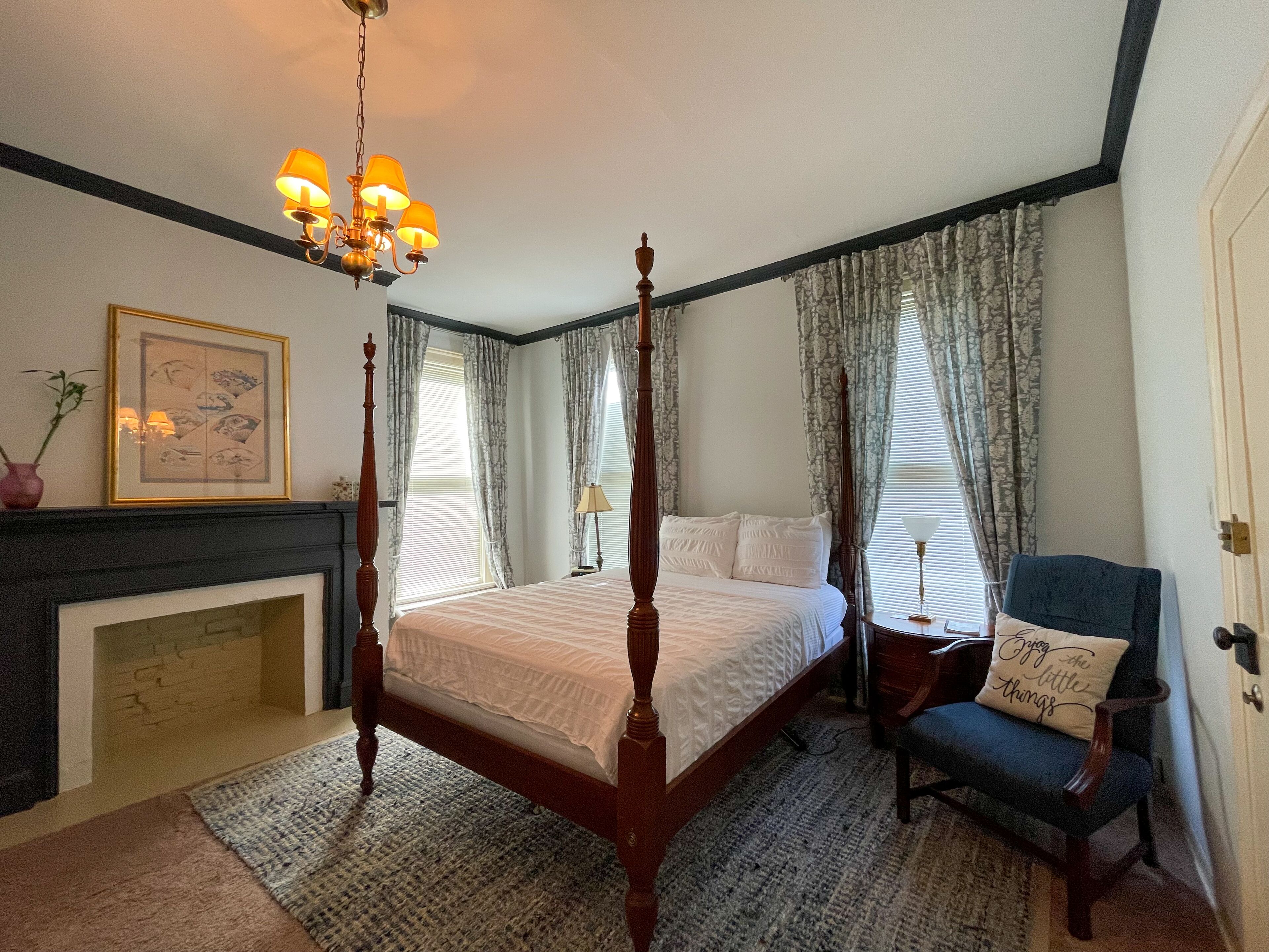 Un lit à baldaquin avec une couvre-lit blanche, une cheminée, un tableau encadré au mur et un fauteuil avec un coussin.