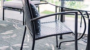 A covered patio area with a glass table and metal chairs, a cushioned bench, and a view of greenery and red umbrellas in the background. - Meadows Inn Bed & Breakfast (New Bern)