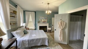 A bedroom with a bed, a bedside table, a chair, a fireplace, a robe hanging on the door, and a bookshelf. - Meadows Inn Bed & Breakfast (New Bern)