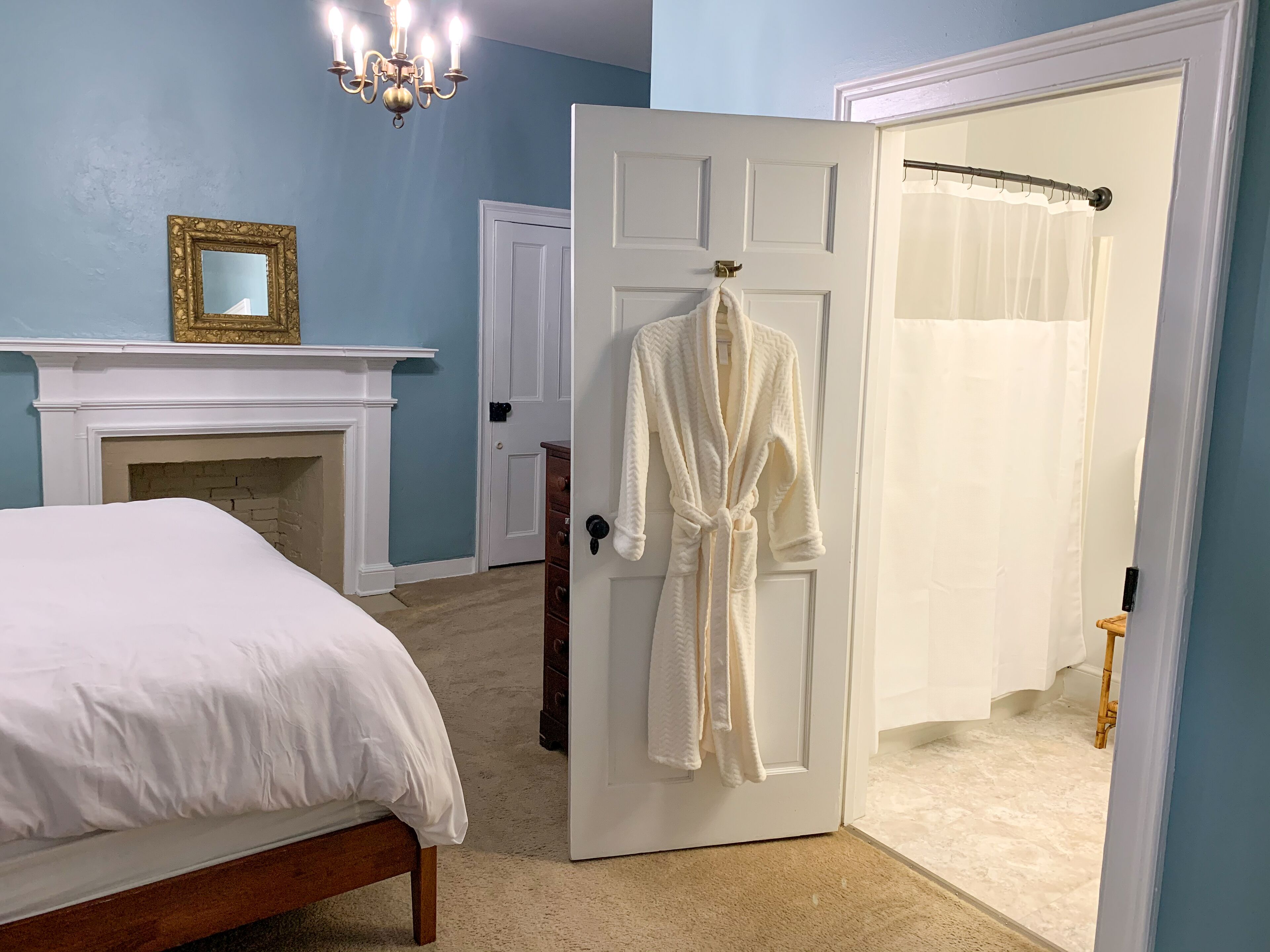 A bedroom with a bed, a white robe hanging on a door, a fireplace, and a chandelier.