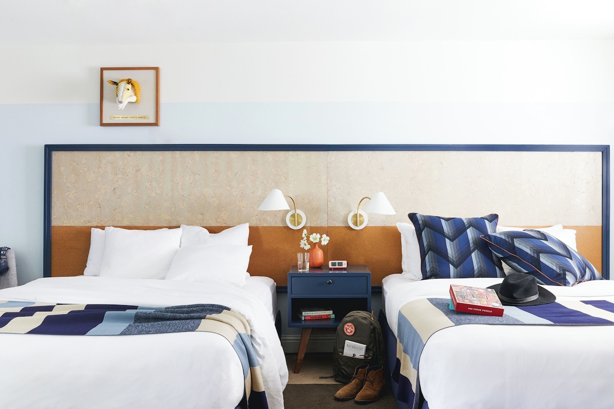 A hotel room with two beds, a nightstand with a lamp, a framed picture, and a small table with a vase and books.