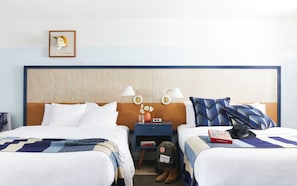 Quarto de hotel com duas camas, uma mesa de cabeceira com abajur, um quadro emoldurado e uma mesinha com um vaso e livros.