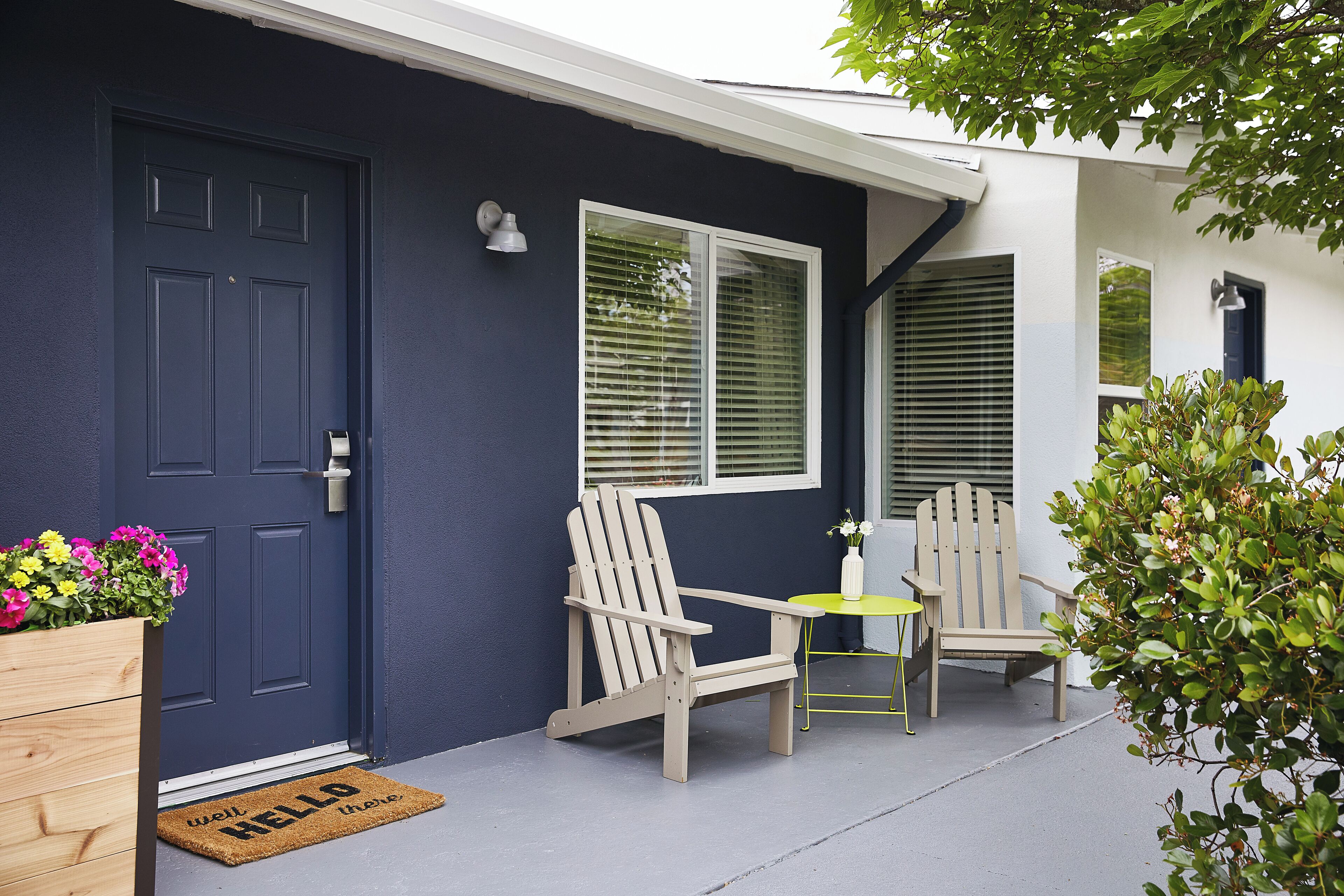 Una casa azul oscuro con puerta de madera, dos sillas Adirondack, una mesita y una planta en maceta.