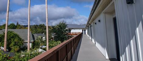A corridor with a wooden railing, leading to a building with a white exterior and a dark door.