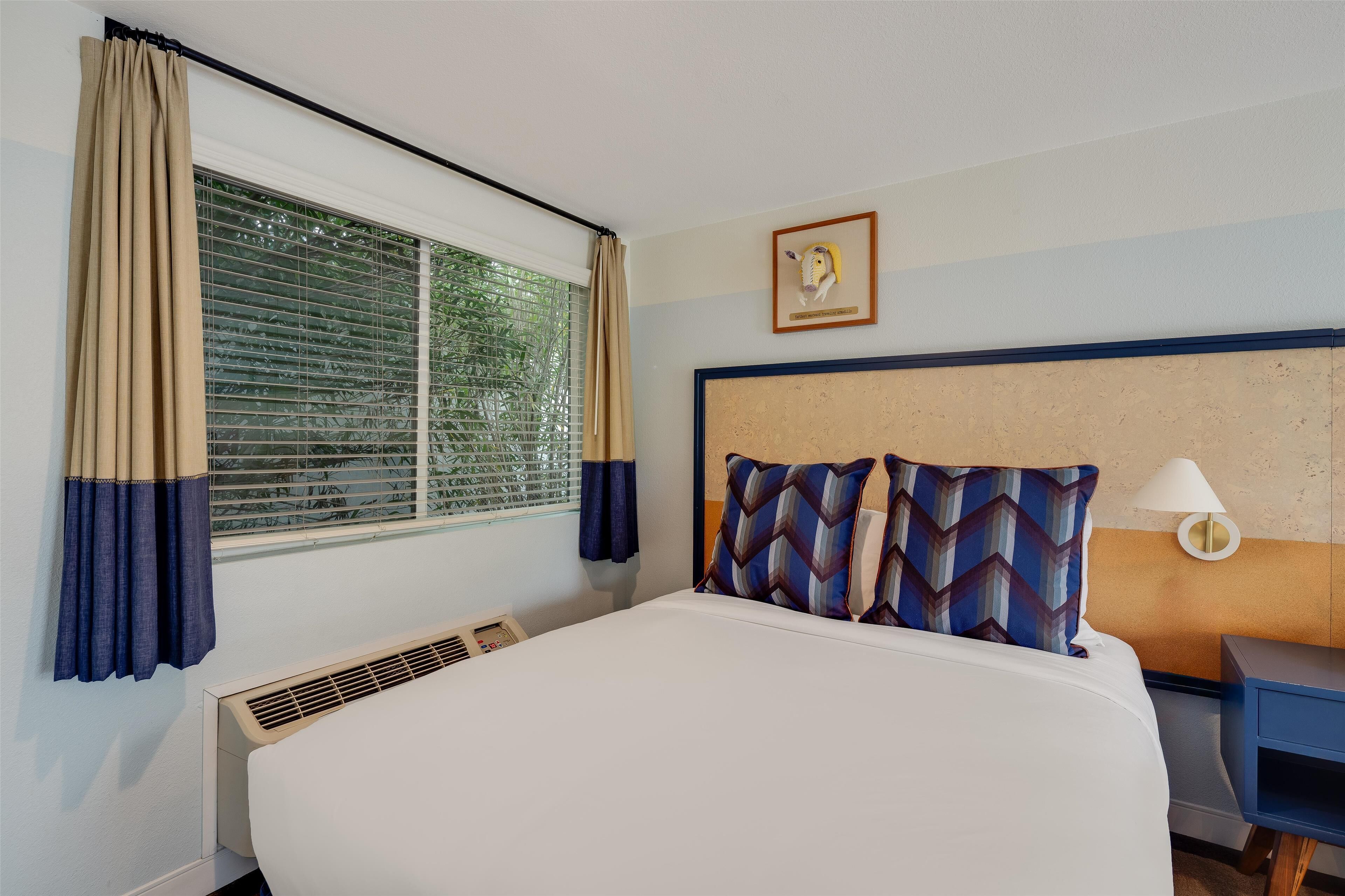 A hotel room with a large bed, a window with blinds, a wall-mounted picture, a bedside table with a lamp, and a wall heater.