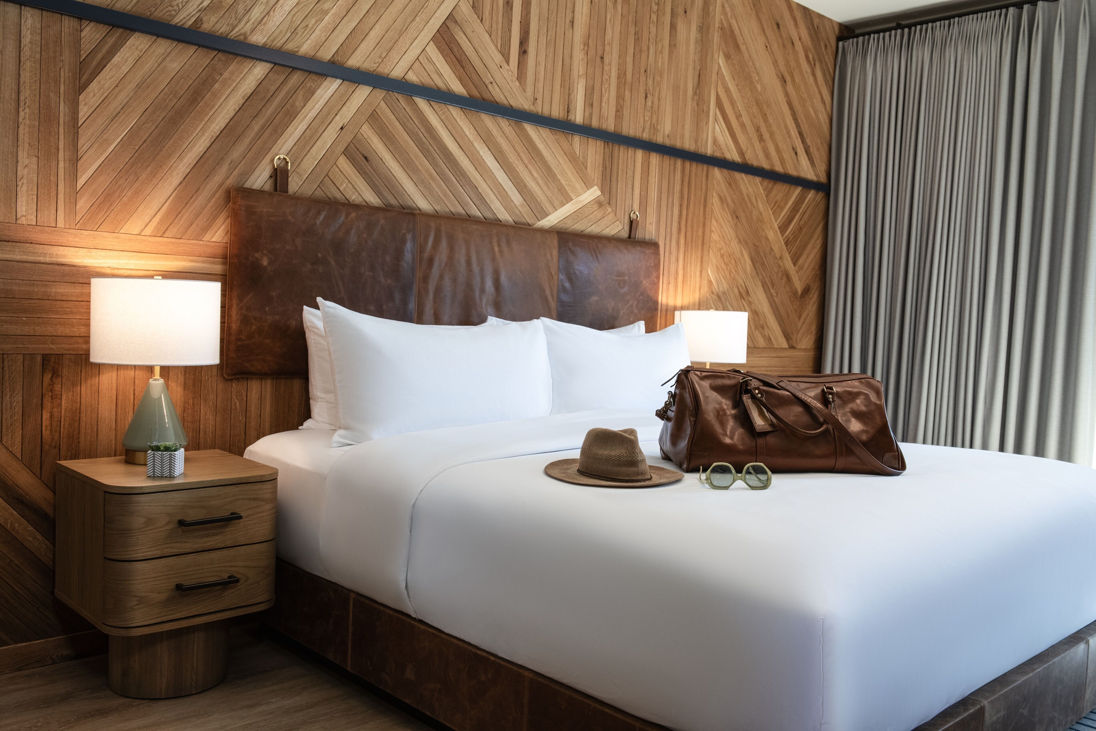 A neatly made bed with a wooden headboard, a bedside lamp, a hat, and a handbag on the bed.