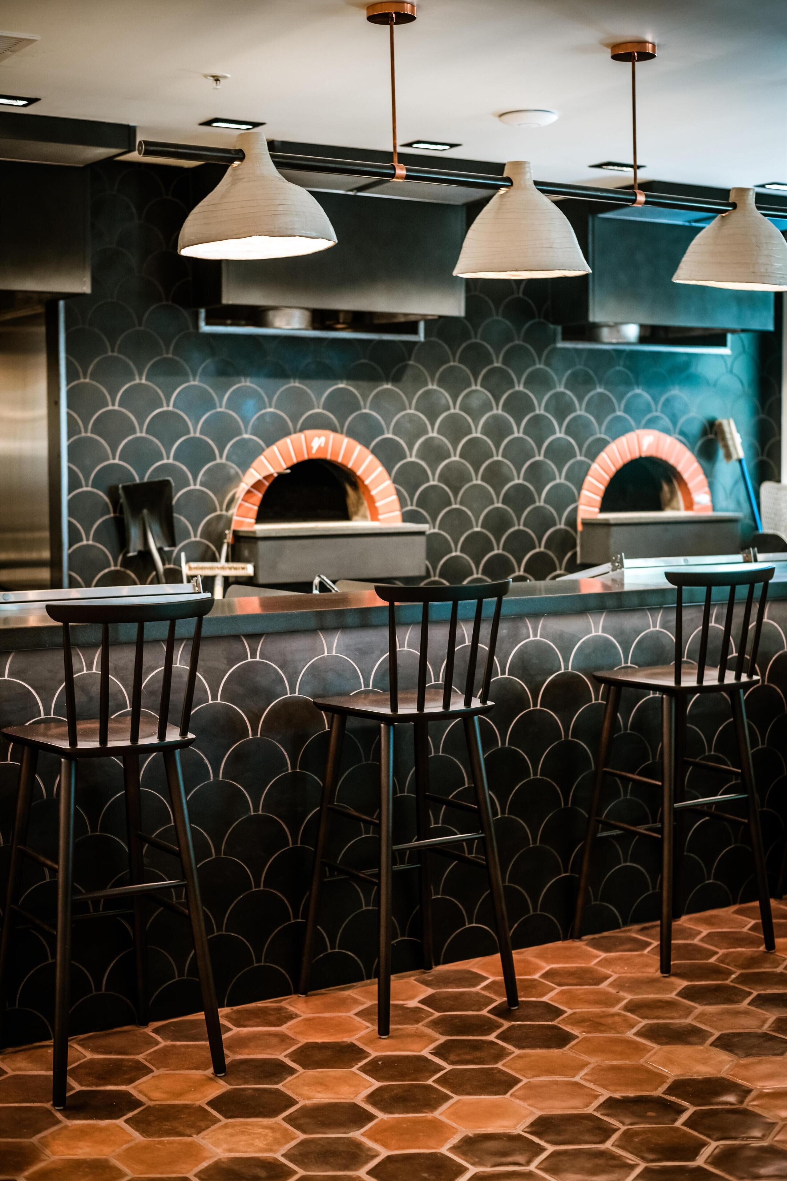 A bar area with a hexagonal patterned floor, black bar stools, and a brick oven in the background.