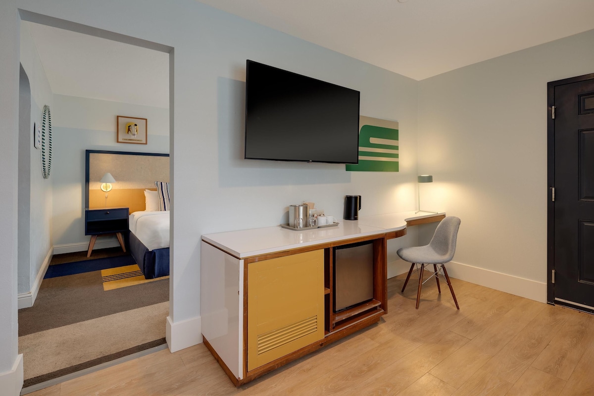 A modern hotel room with a flat-screen TV mounted on the wall, a small desk with a chair, a bed with a blue headboard, and a wooden cabinet with a microwave.