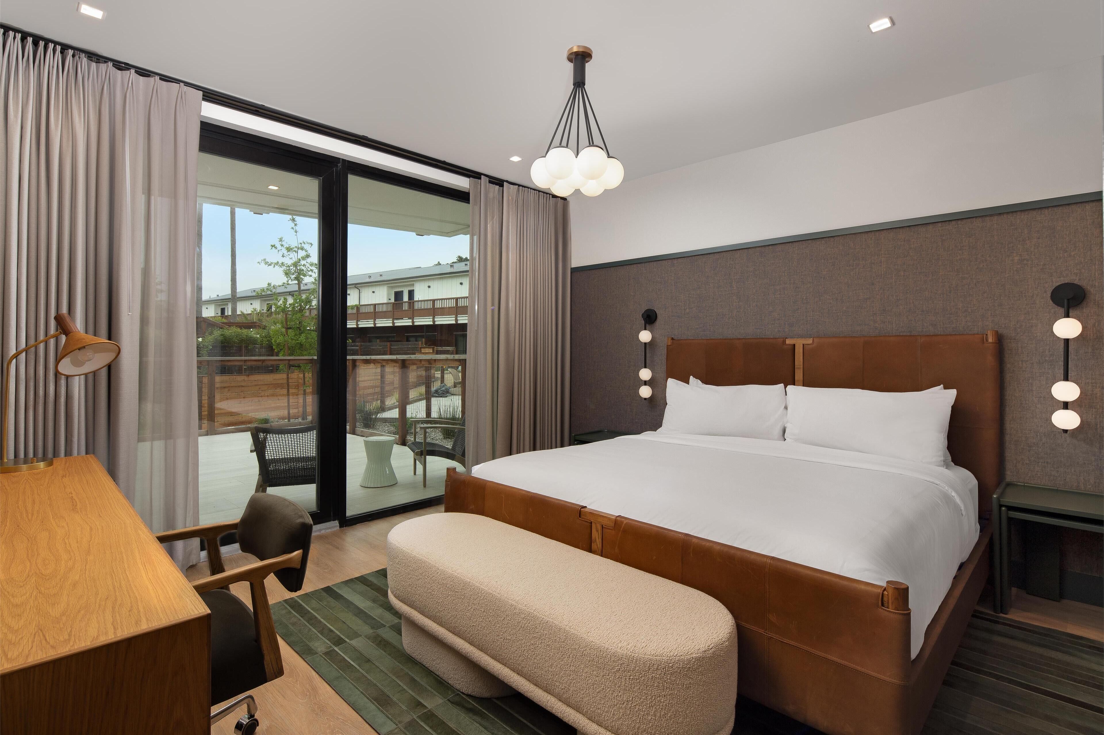 A hotel room with a large bed, a desk, a chair, a bench, and a view of the outside.