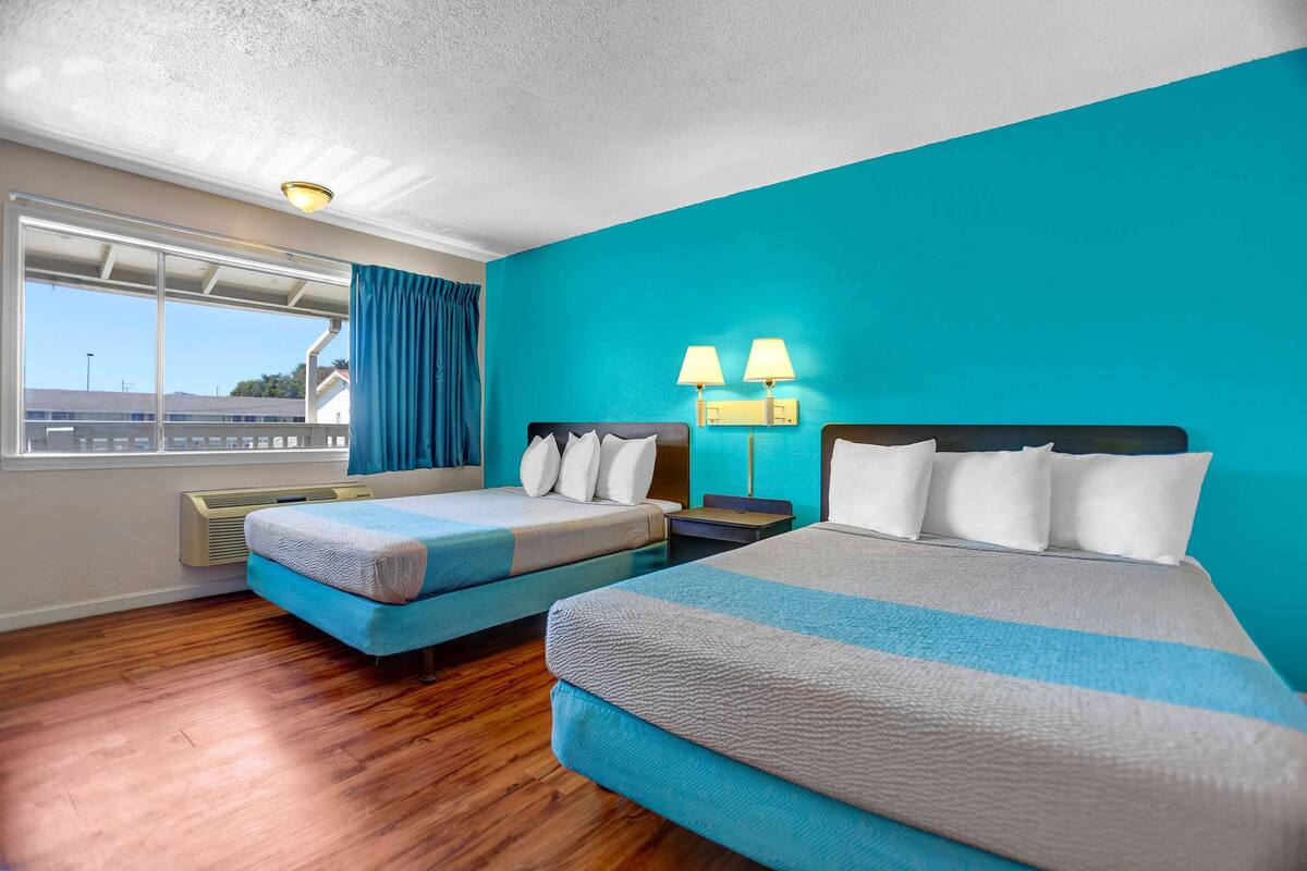 A hotel room with two beds, a large window with curtains, and a view of the outside.
