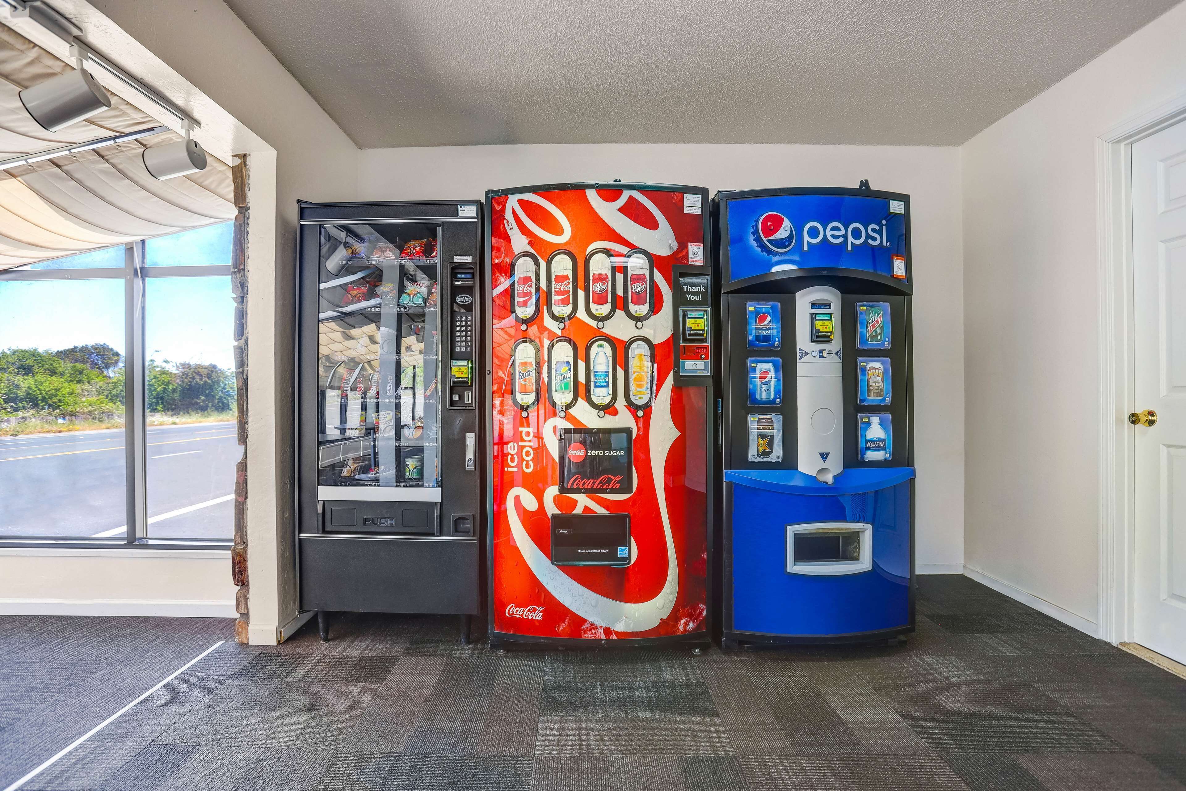 Three beverage vending machines in a room with a large window and carpeted floor.