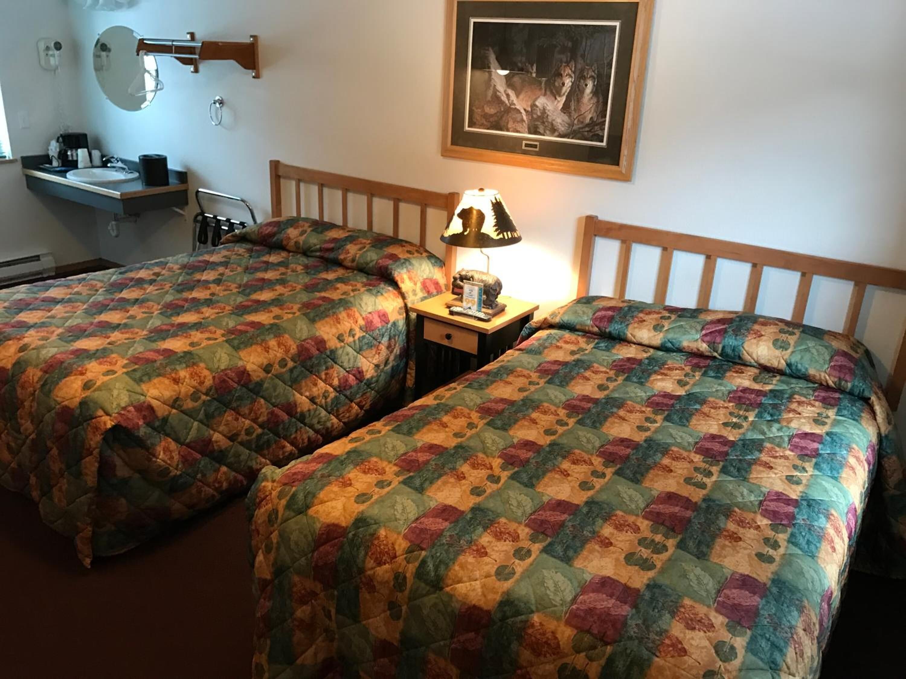 Une chambre d’hôtel avec deux lits, une table de chevet avec une lampe, un miroir, un tableau encadré au mur et un petit bureau avec un lavabo.