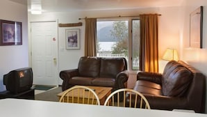 A living room with a brown leather sofa, a wooden coffee table, a television, and a window with curtains. - Harborview Inn (Seward)