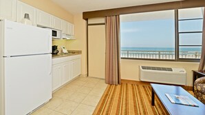 A compact kitchen with a refrigerator, microwave, and a coffee maker. There's a view of the ocean through a window, and a table with a magazine on it.