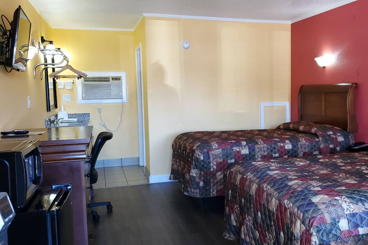 A hotel room with two beds, a desk with a chair, a desk lamp, an air conditioning unit, and a wall-mounted light fixture.