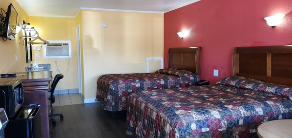 A hotel room with two beds, a desk with a chair, a desk lamp, an air conditioning unit, and a wall-mounted light fixture.