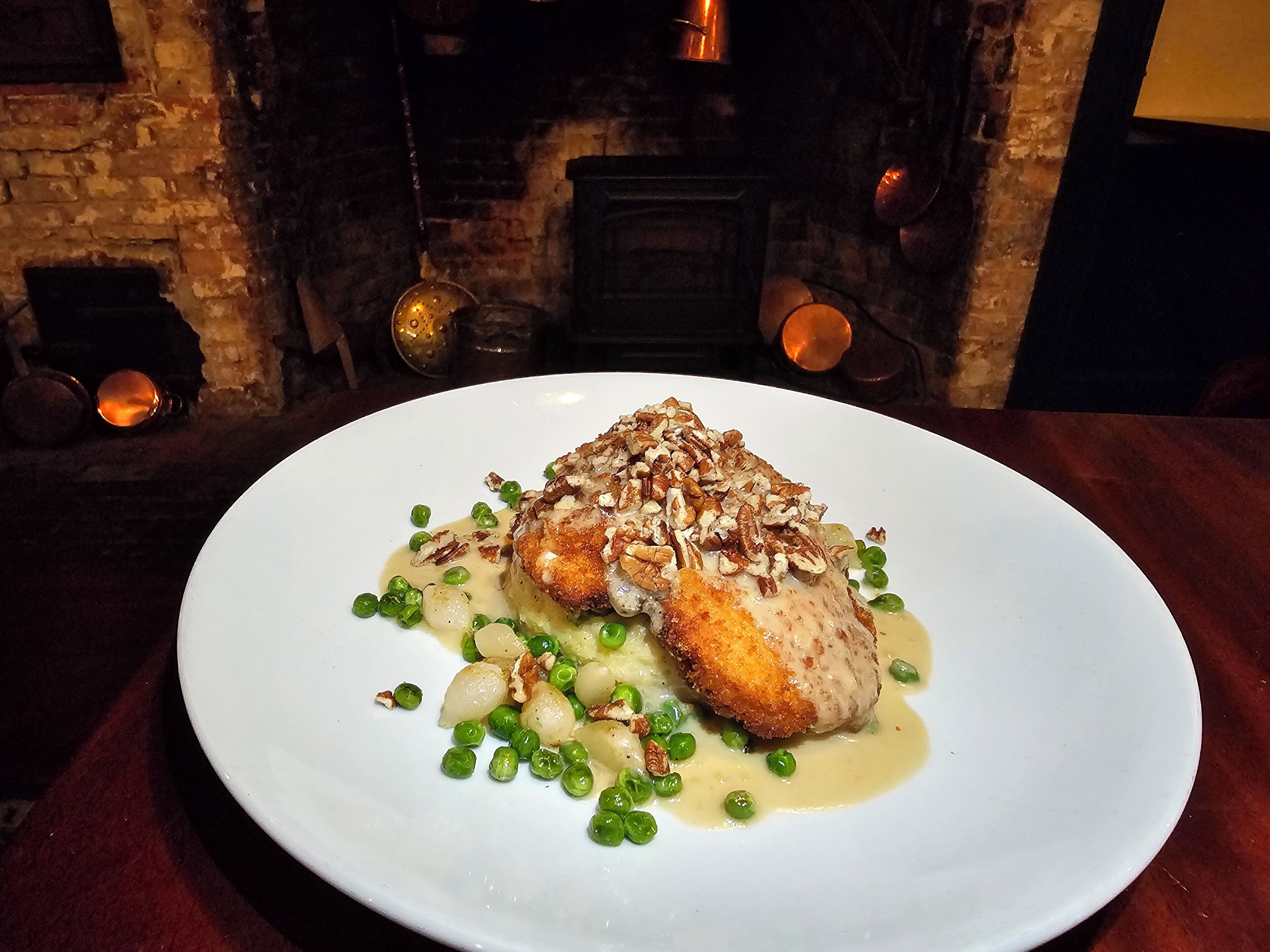 A white plate with a piece of food, green peas, and a sauce, set on a table with a brick wall and copper pots in the background.