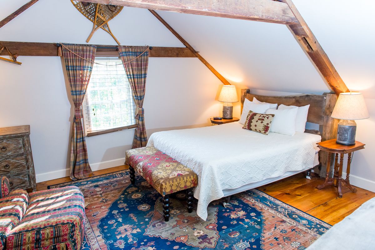 A cozy attic bedroom with a wooden ceiling, a large bed with white linens, two bedside lamps, a bench, and a patterned rug.