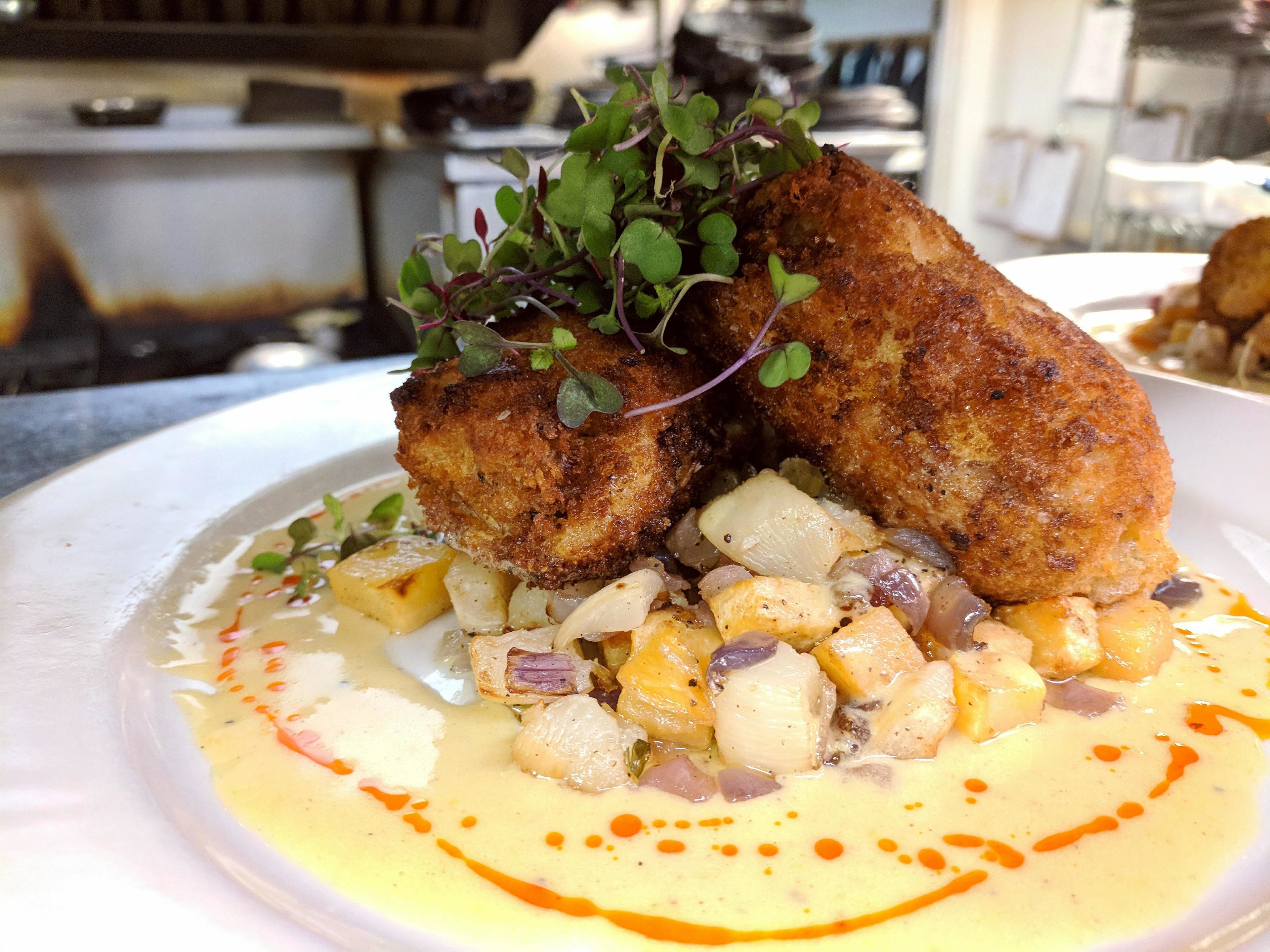 A plated dish with a golden-brown fried item, diced potatoes, and a creamy sauce with orange drizzle.