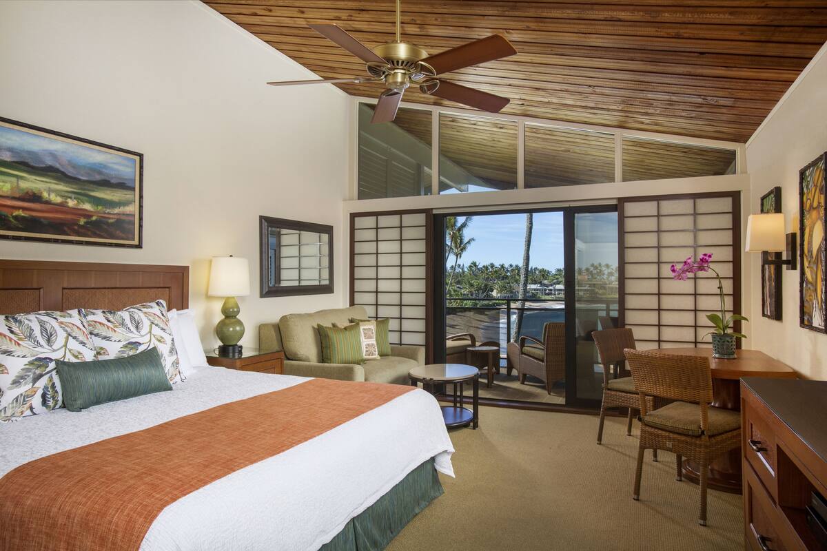 Quarto de hotel com uma cama grande, um ventilador de teto, uma varanda com vista para o oceano e portas deslizantes japonesas tradicionais.