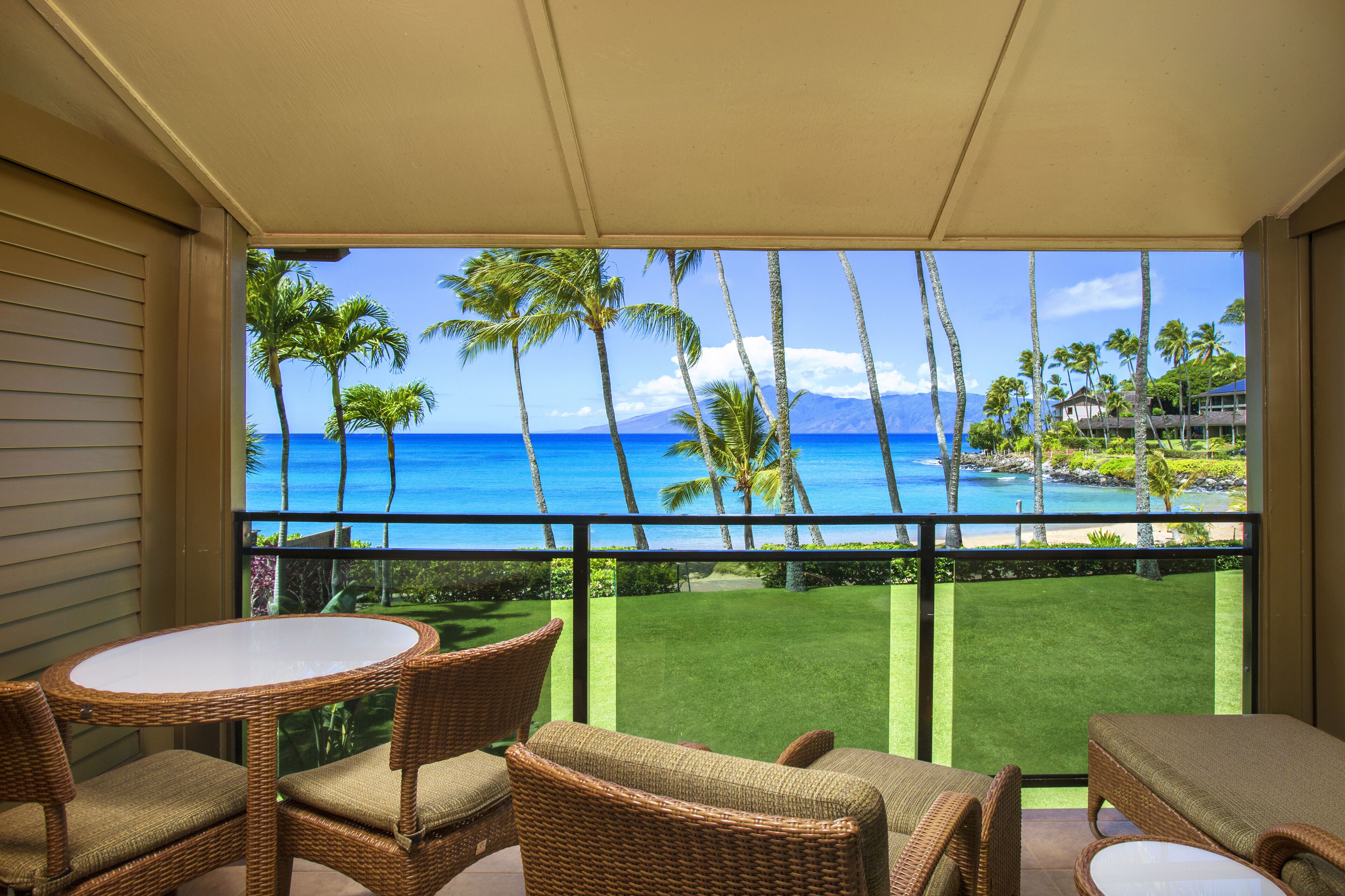 A balcony with wicker furniture, a view of the ocean, palm trees, and a golf course.