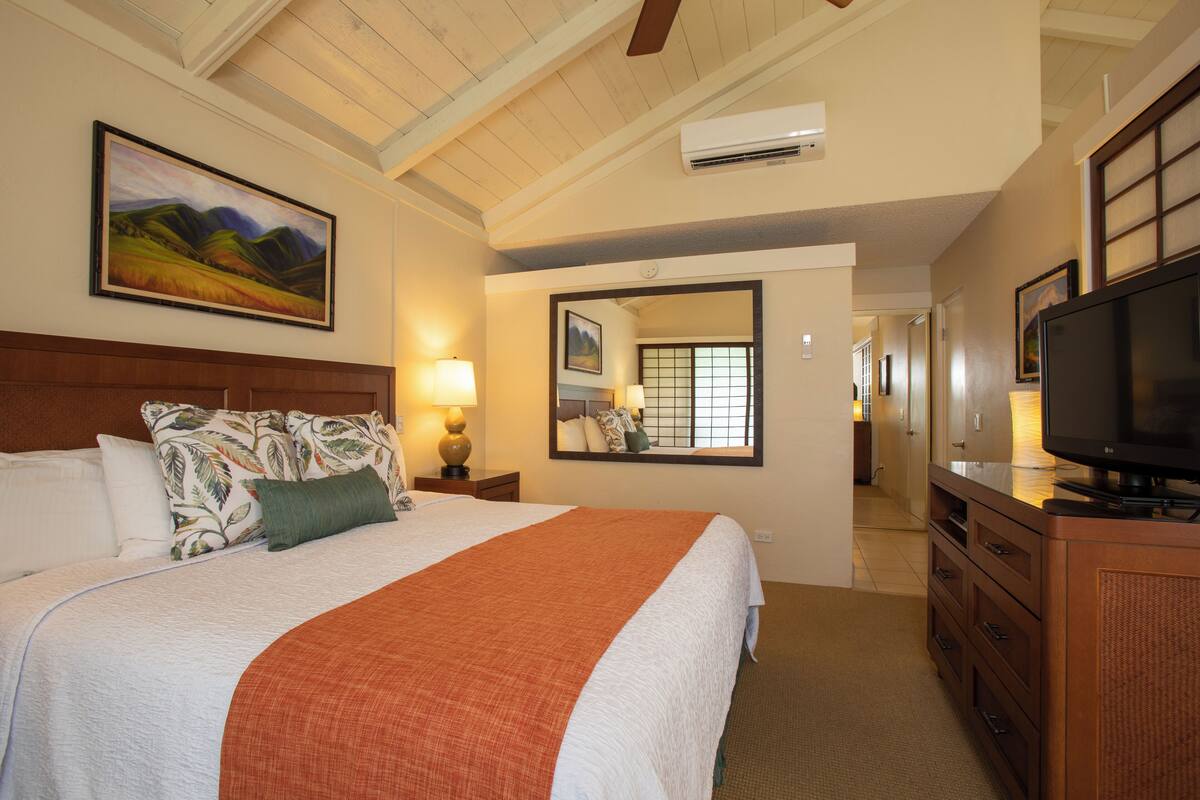 Quarto de hotel com uma cama grande, televisão, cômoda e corredor que dá acesso a outros quartos.